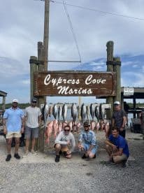 Group of anglers proudly posing in front of a catch board displaying a variety of fish, including Snapper and Tuna, at Cypress Cove Marina.