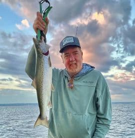 An angler proudly displaying a large Speckled Trout while fishing on a calm lake, with a beautiful sunset backdrop.