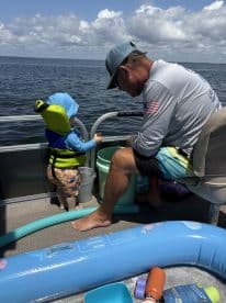 A young child in a life jacket observes an adult on a pontoon boat, enjoying a sunny day on the water.