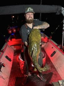 Angler proudly displaying a large Catfish while fishing at night, showcasing the vibrant atmosphere of the boat with red lighting.