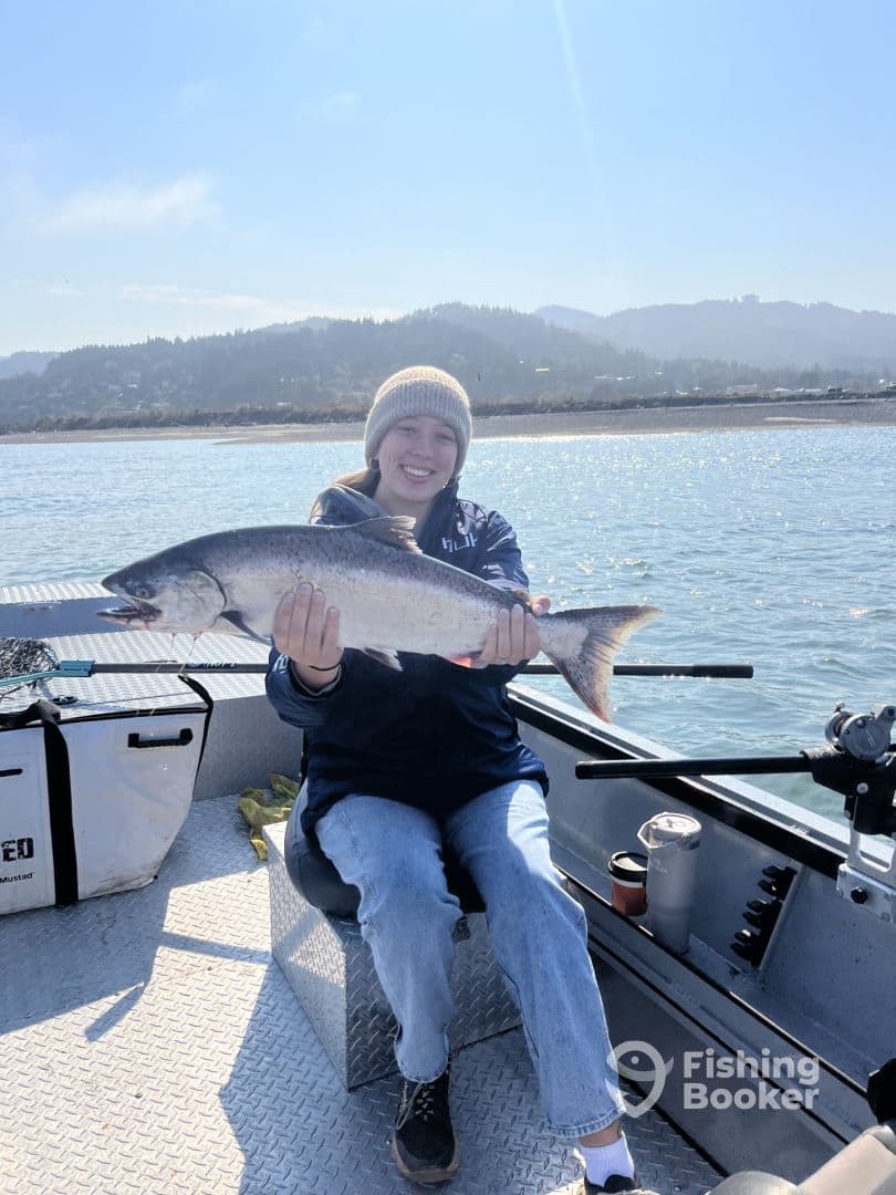 Angler proudly displaying a large salmon while seated on a fishing boat, with a scenic backdrop of hills and water.