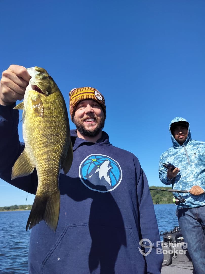 Angler proudly displaying a large Smallmouth Bass while fishing on a serene lake, with another fisherman in the background.