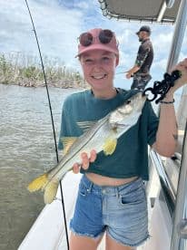 A young woman proudly displaying a Snook caught while fishing on a boat in a scenic waterway.
