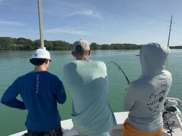 Three anglers engaged in fishing on a boat, with one actively reeling in a catch in a scenic waterway.