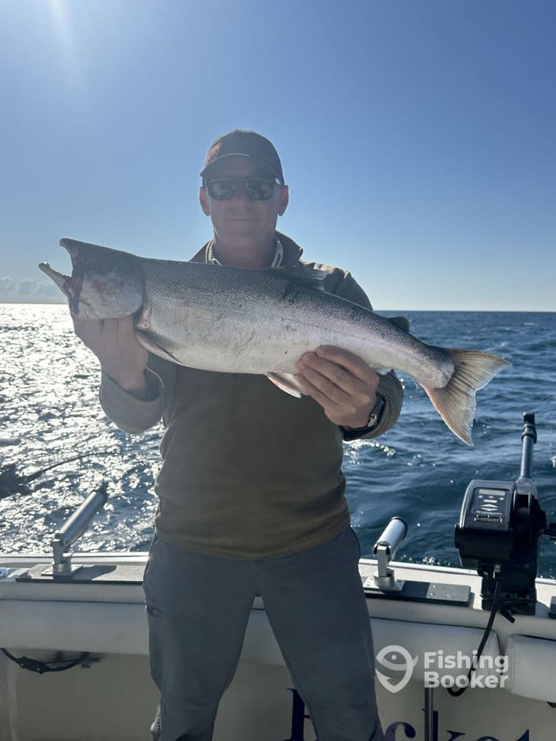 An angler proudly displaying a large Lake Trout while fishing on a sunny day, with the shimmering water in the background.