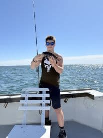 A young angler proudly displaying a freshly caught Black Sea Bass while fishing on a boat in clear waters.
