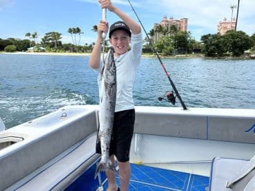 A young angler proudly displaying a large Barracuda while fishing from a boat in a tropical setting.