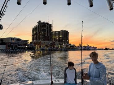Two young anglers enjoying a sunset fishing trip on a boat, with a vibrant waterfront backdrop and fishing rods ready for action.