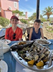 Two young boys enjoying a meal featuring grilled fish and garnishes at a seaside restaurant, showcasing a relaxed dining experience after a fishing trip.