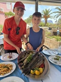 Two young boys proudly presenting a platter of grilled fish, garnished with lemon and herbs, at a seaside restaurant.