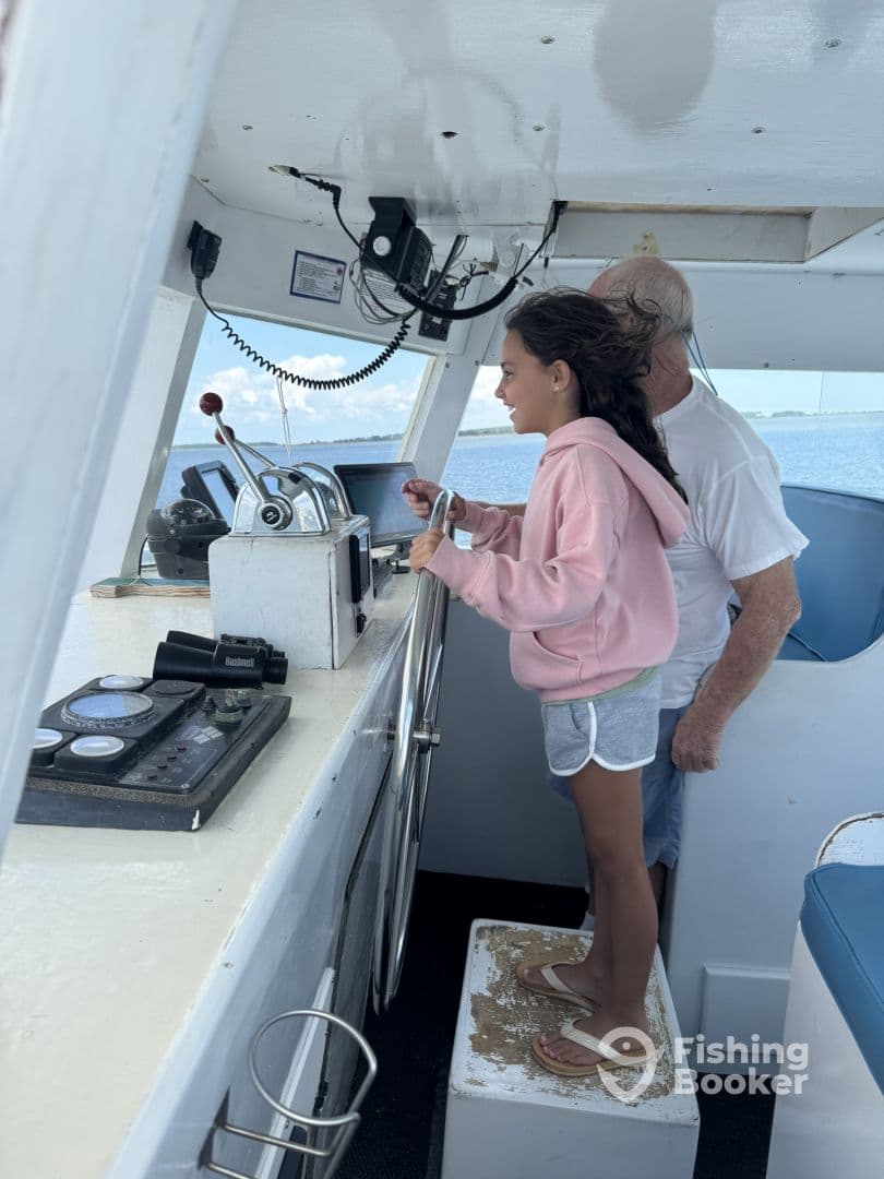 A young girl is at the helm of a fishing boat, learning to steer with guidance from an adult, showcasing a fun and educational fishing experience.
