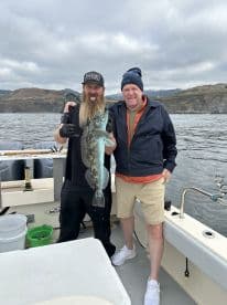 Two anglers proudly displaying a large Lingcod on a fishing boat, showcasing a successful day on the water.