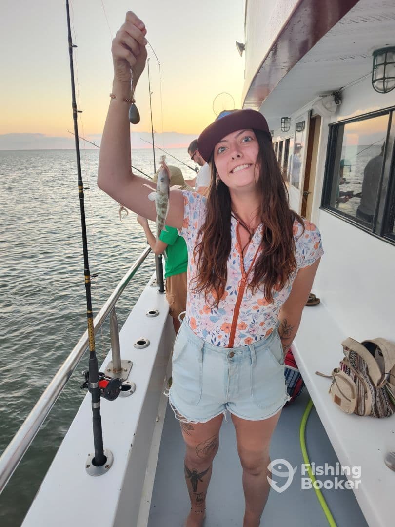An enthusiastic angler proudly displaying a small fish while fishing on a boat during sunset.