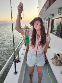 An enthusiastic angler proudly displaying a small fish while fishing on a boat during sunset.