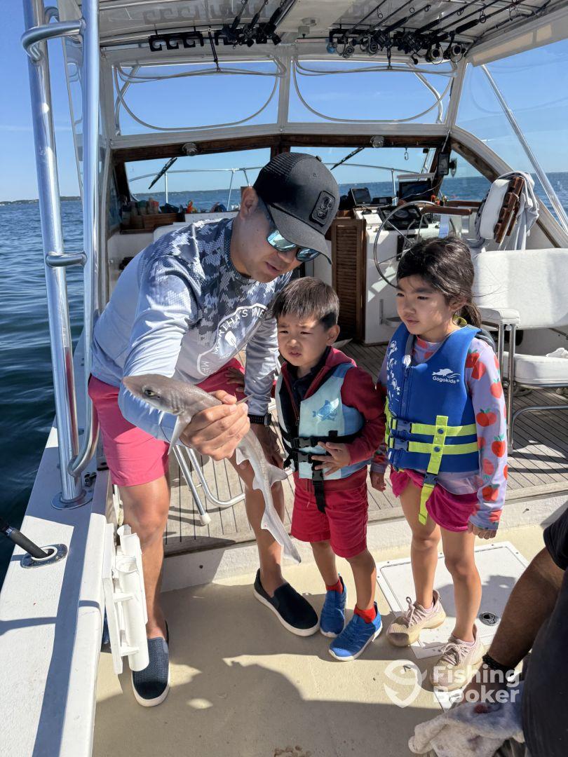 A young boy and girl, both wearing life jackets, watch excitedly as an adult holds a small shark on a fishing boat, showcasing a family fishing experience.