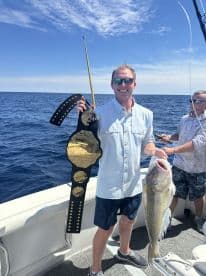 Angler proudly displaying a large striped bass while holding a championship belt on a fishing charter in open waters.