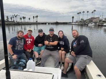 A group of five anglers enjoying a day on the water, seated inside a boat with a scenic harbor view in the background.