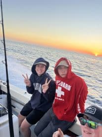 Three young anglers enjoying a sunset on a fishing boat, showcasing a relaxed atmosphere with the ocean in the background.
