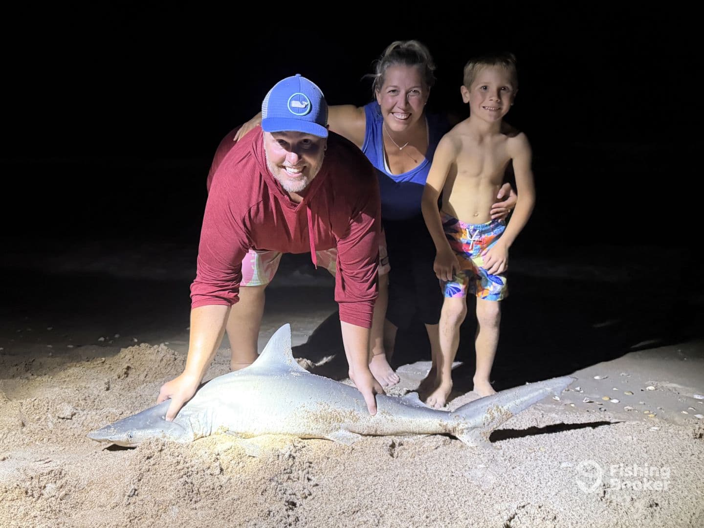 A family proudly displaying a large shark caught at night on the beach, showcasing a memorable fishing experience.