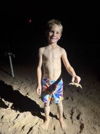 A young boy proudly displaying a crab while standing on the beach at night, showcasing a fun family fishing experience.
