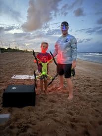 A man and a young boy are standing on the beach at dusk, holding a drone, with no visible fishing activity or catch.