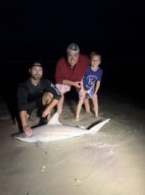 A family poses proudly with a caught shark on the beach at night, showcasing their fishing success.