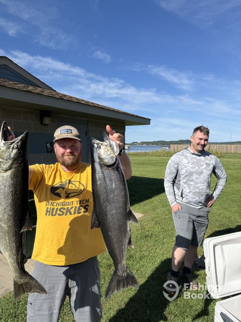 Two anglers proudly displaying their catch of large Salmon in front of a lakeside building after a successful fishing trip.