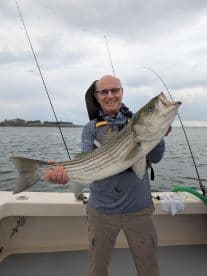 An angler proudly displaying a large Striped Bass while fishing from a boat, showcasing a successful day on the water.