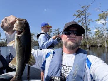 An angler proudly displaying a large Largemouth Bass while fishing in a scenic freshwater environment, with another fisherman in the background.