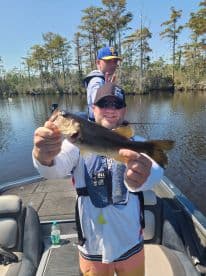 An angler proudly displaying a Largemouth Bass while fishing in a scenic freshwater environment, with a young companion in the background.