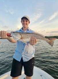 A young angler proudly displaying a large Redfish while fishing on a boat during sunset.