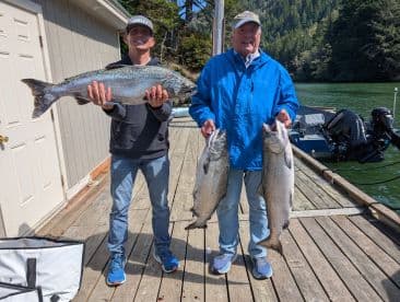 Two anglers proudly displaying their catch of Salmon at the dock, showcasing a successful day of fishing in a scenic coastal environment.
