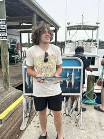 A young angler proudly displaying a Spanish Mackerel at the dock after a fishing trip, with multiple fish hanging in the background.