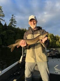 An adult angler proudly displaying a Northern Pike while fishing on a serene lake, surrounded by lush greenery.