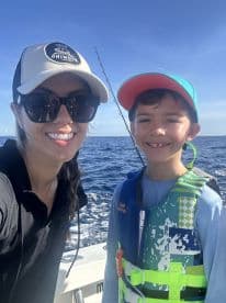 A smiling woman and a young boy wearing life jackets pose for a selfie on a boat, with the ocean in the background.