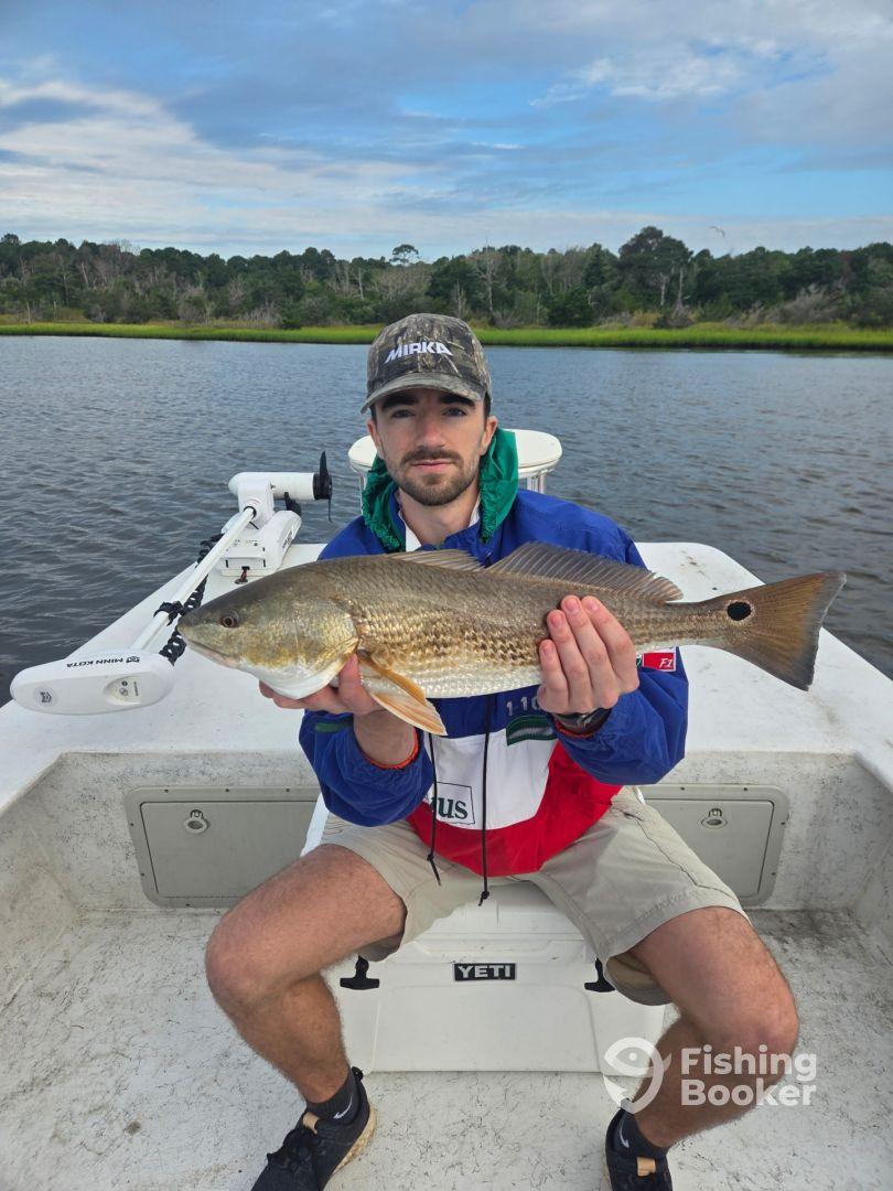 Angler holding a Redfish on a boat in a scenic waterway, showcasing a successful catch during a fishing trip.