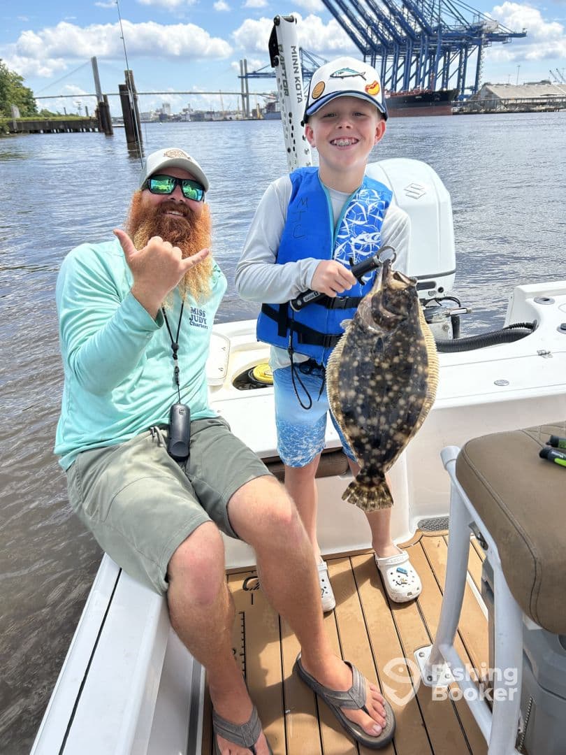A young angler proudly displaying a Flounder while fishing on a boat, accompanied by an adult. The backdrop features a scenic waterway and industrial structures.