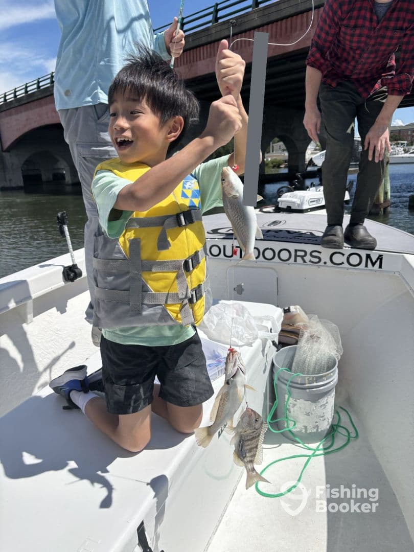 A young angler in a life jacket joyfully holds up two fish while kneeling on a boat, showcasing a successful fishing trip.