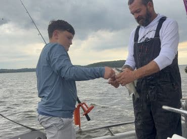 A young angler and an adult are engaged in a fishing moment, with the boy holding a freshly caught fish on a boat in a scenic waterway.