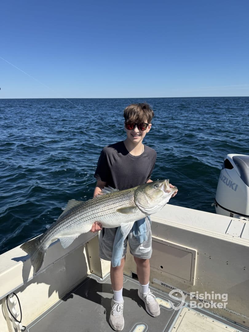 A young angler proudly displaying a large Striped Bass while fishing on a boat in clear waters.