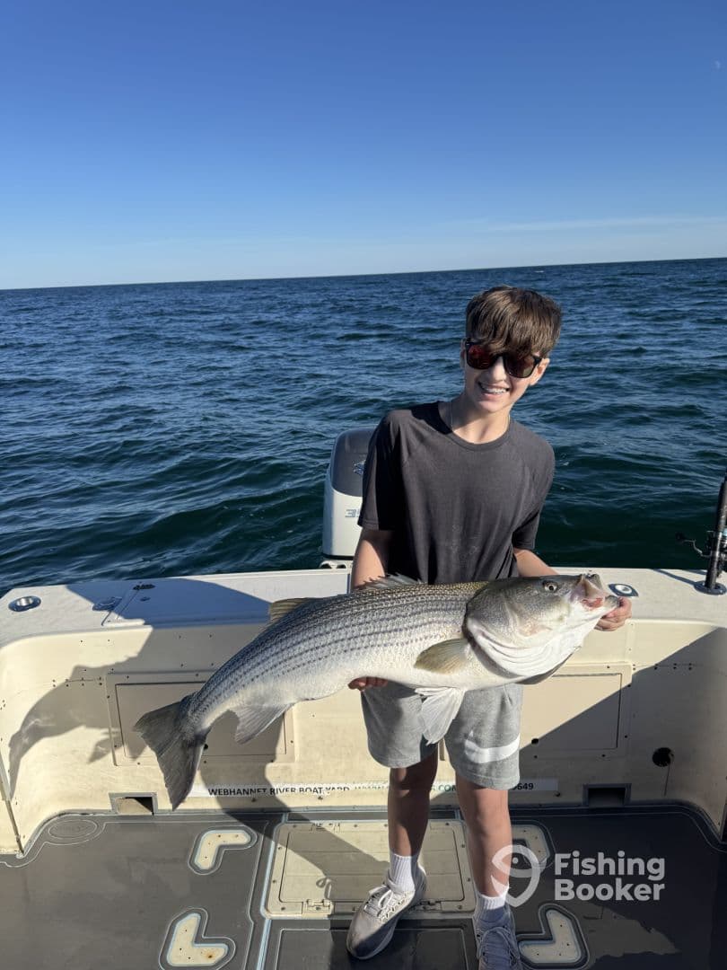 A young angler proudly displaying a large Striped Bass while fishing on a boat in open waters.
