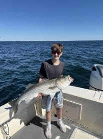 A young angler proudly displaying a large Striped Bass while fishing on a boat in clear waters.