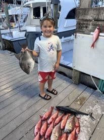 A young angler proudly displaying a large Flounder at the marina, surrounded by a variety of freshly caught fish after a successful fishing trip.