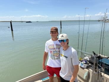 Two young anglers posing on the deck of a fishing boat, with a scenic view of the calm waters and distant boats in the background.