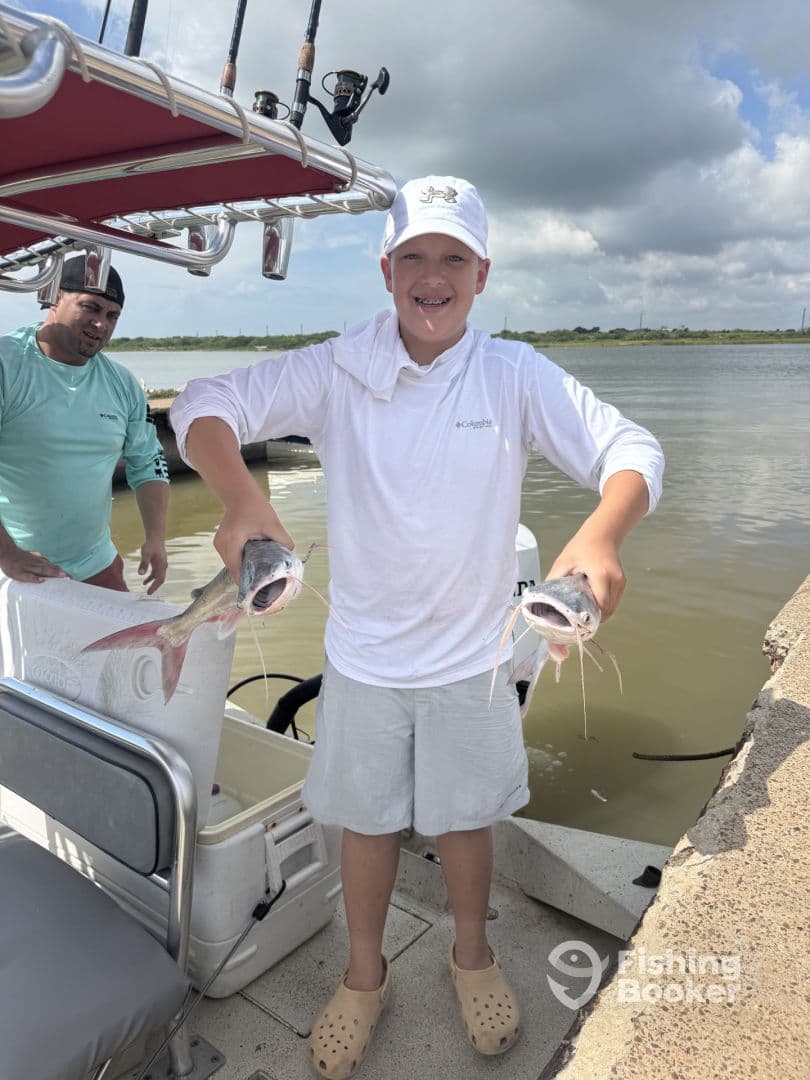 A young angler proudly displaying two Catfish while standing on a boat dock, showcasing a successful fishing trip.