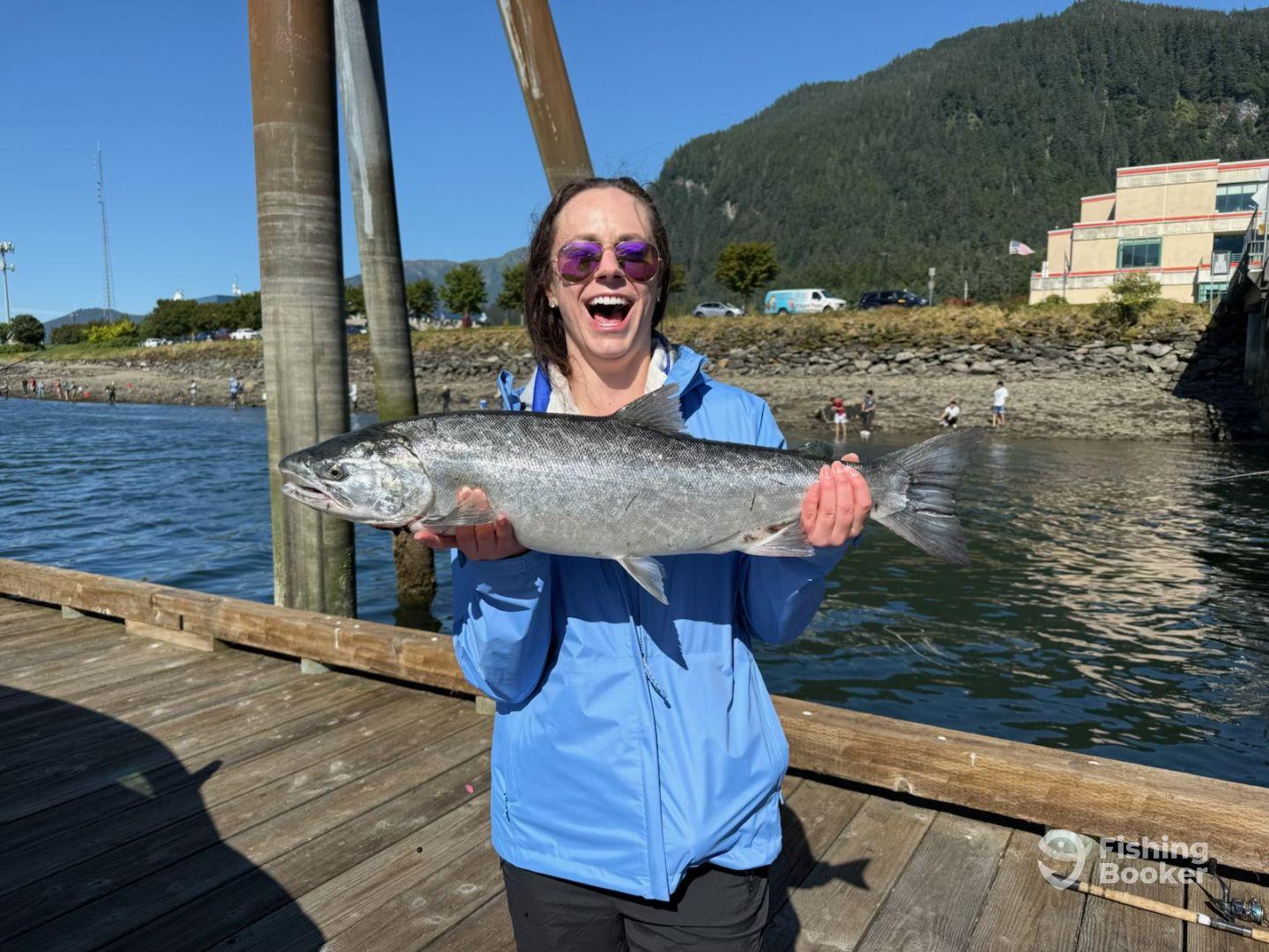 A joyful angler proudly displaying a large salmon at the dock, surrounded by a scenic waterfront and distant fishing activity.
