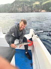 Angler holding a Barracuda on a fishing boat, surrounded by scenic coastal cliffs, showcasing a successful fishing trip.