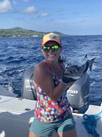 A smiling angler proudly displays a large Barracuda while fishing off the coast, showcasing the vibrant blue waters and lush green hills in the background.