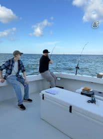 Two anglers engaged in fishing on a charter boat, with one actively reeling in a line while the other observes, set against a scenic ocean backdrop.