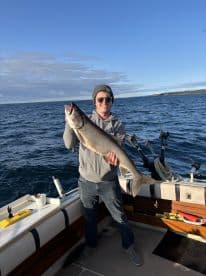 Angler proudly displaying a large Lake Trout while fishing on a boat in a scenic waterway.